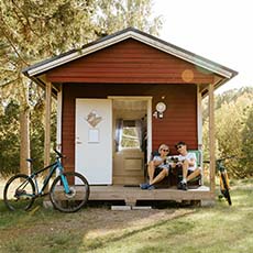 En rödstuga där ett par sitter och läser på verandan. Motknuten lutar en cykel. Foto Henrik Mill.