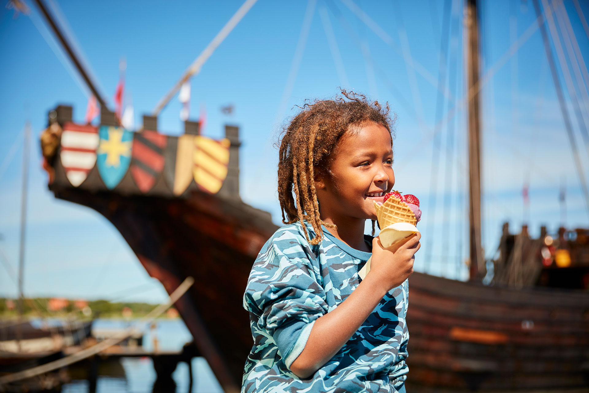 Ett barn äter en glass på Frösåkers Brygga i Västerås med ett stort vikingaskepp i bakgrunden. Foto: BildN