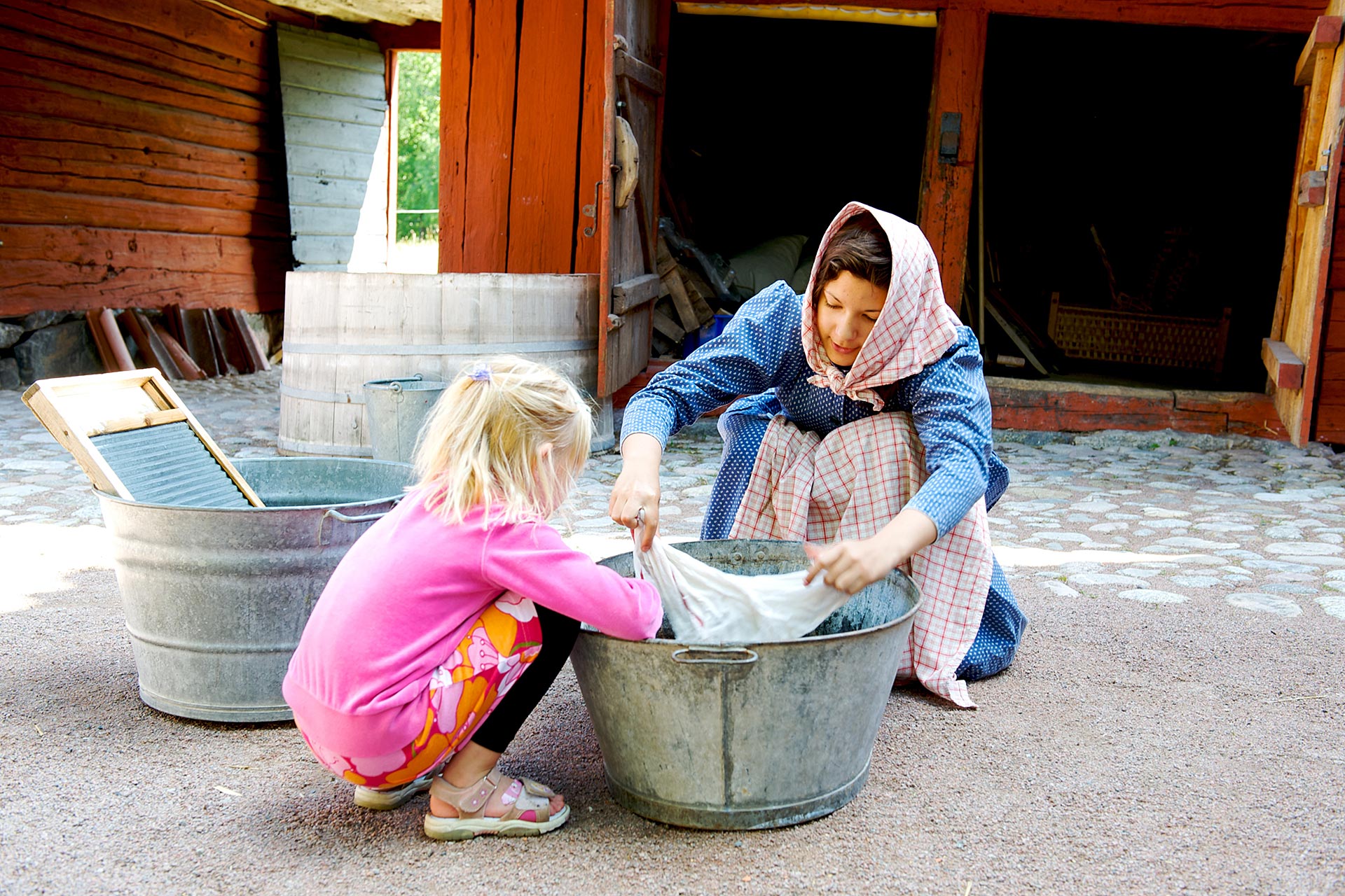 En kvinna från Vallby Fridluftsmuseum i Västerås utklädd till piga tvättar tvätt i tvättbalja med ett barn som är på besök. Fotograf: Leon Grimaldi