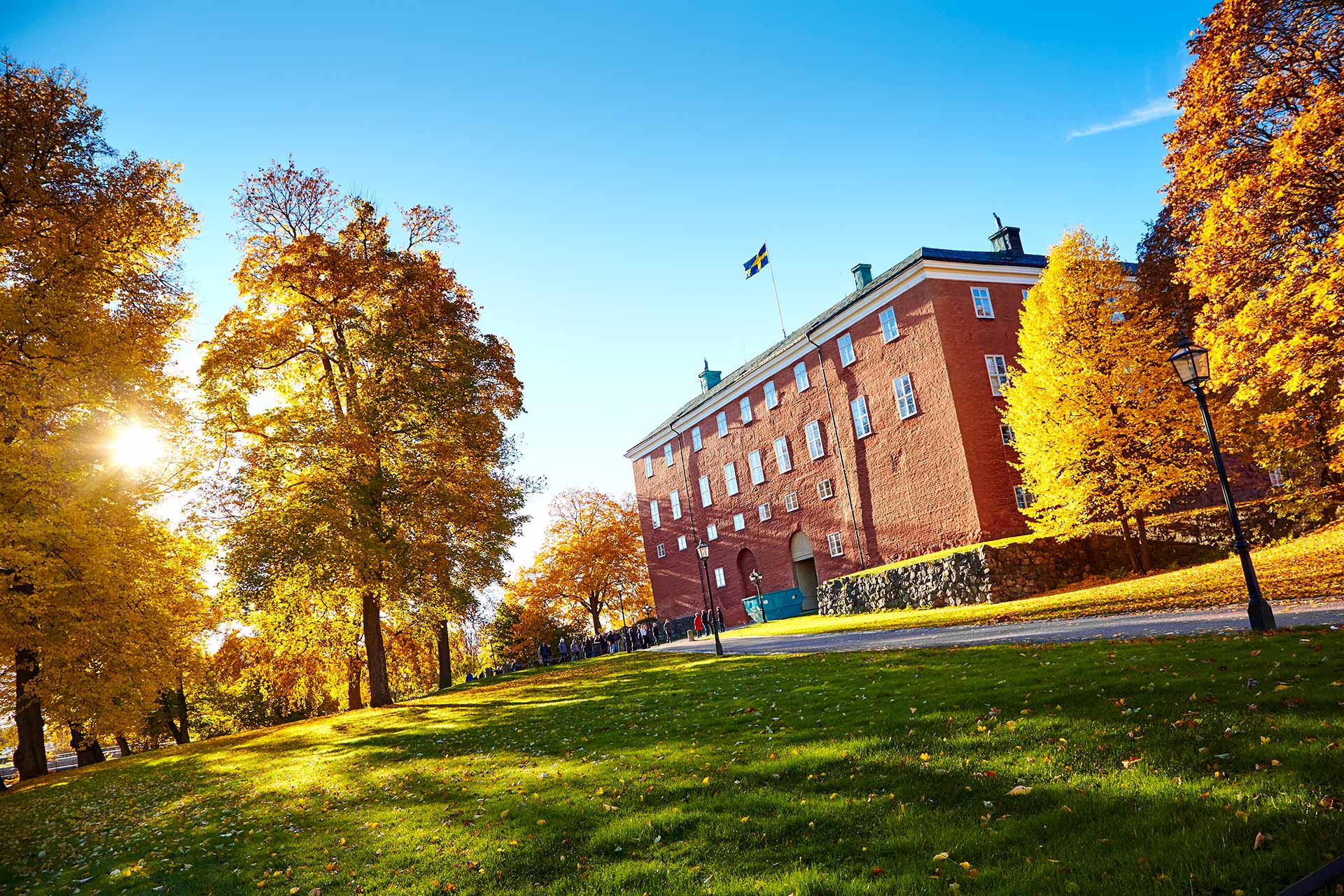Västerås slott i motljus. Löv i guldfärger pryder träden och en grupp besökare lyssnar på guide. Fotograf: Pia Nordlander